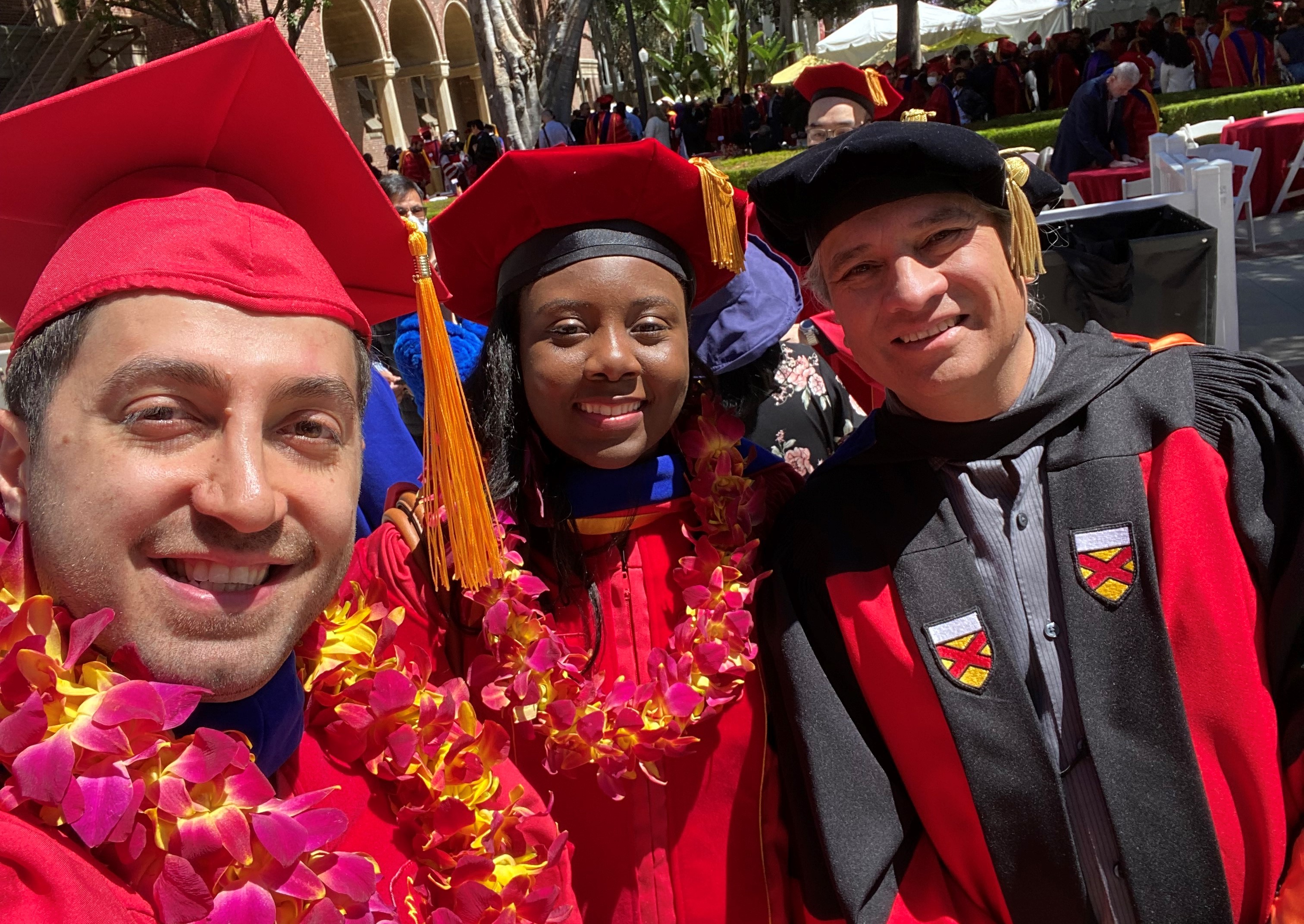 From left to right: Ali Marjaninejad, Jasmine Berry, and Francisco Valero-Cuevas. Ali and Jasmine are wearing red graduation caps and gowns with red and yellow flower leis around their necks. Francisco is wearing a black graduation cap and gown.